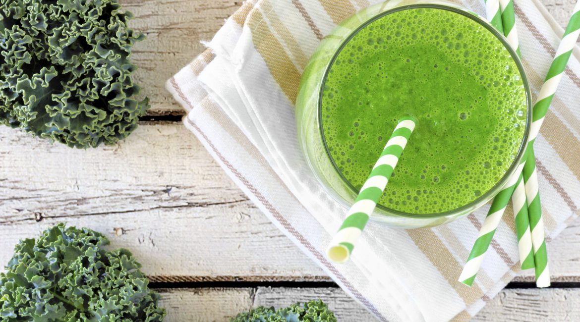Green kale smoothie overhead view, in a glass with straw on a striped cloth against aged wood