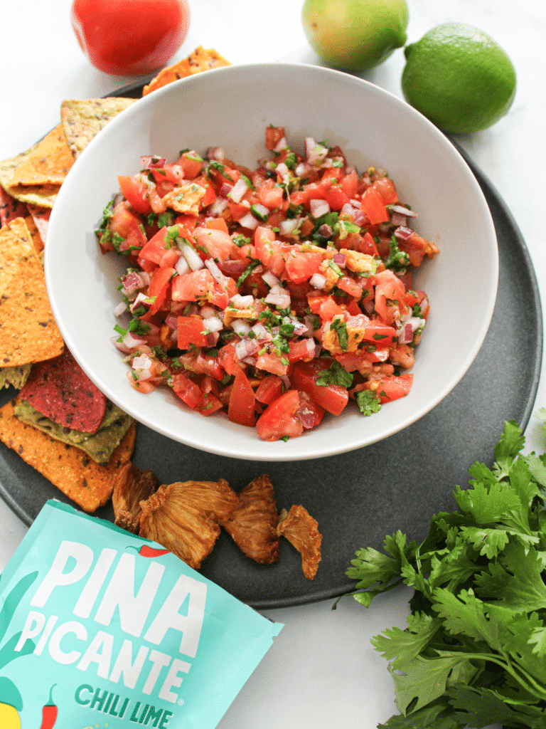 A white bowl full of Pina Picante Chili Lime Pico De Gallo alongside some chips, vegetables, and a bag of Piña Picante Chili Lime.