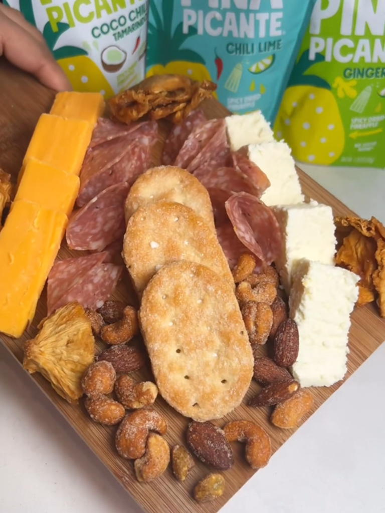 Charcuterie Board that features nuts, salami, crackers, cheeses and all Piña Picante Chili Lime, Ginger Lime, and Coco Chili Tamarindo. Bags of Piña Picante Chili Lime, Coco Chili Tamarindo and Ginger Lime decorate the background.