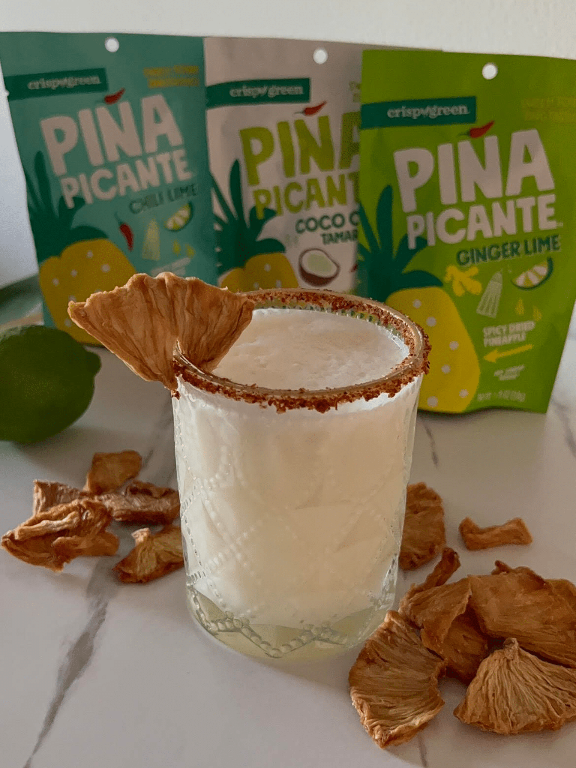 Piña Picante Mocktail glass rimmed with Tajin powder and garnished with a piece of Piña Picante on top of a white table surrounded by other pieces of Piña Picante. The background features bags of Piña Picante Chili Lime, Piña Picante Ginger Lime and Piña Picante Coco Chili Tamarindo.