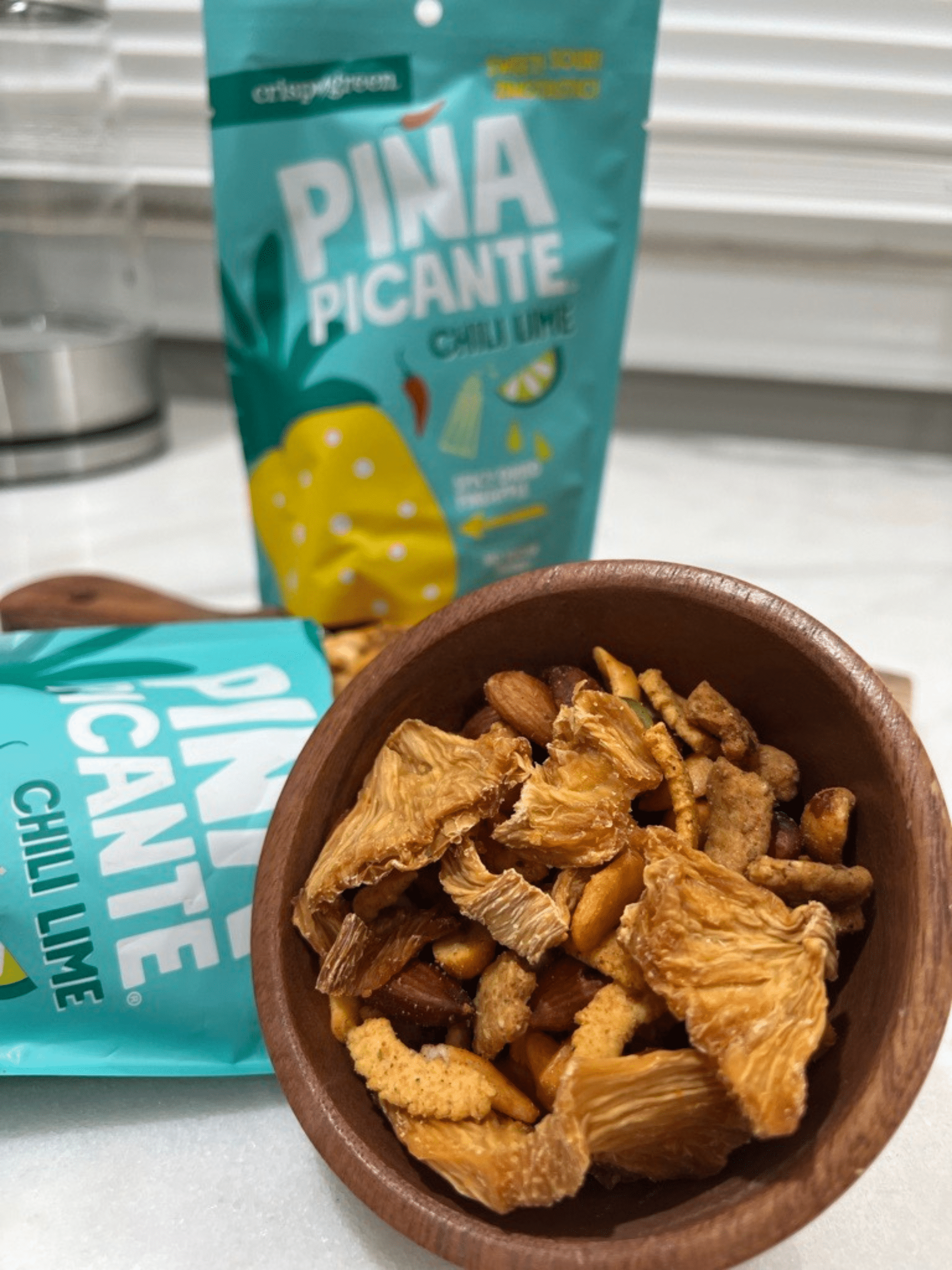 Brown bowl filled with trail mix, including nuts, seeds and pieces of Piña Picante Chili Lime. The background features two bags of Piña Picante Chili Lime.