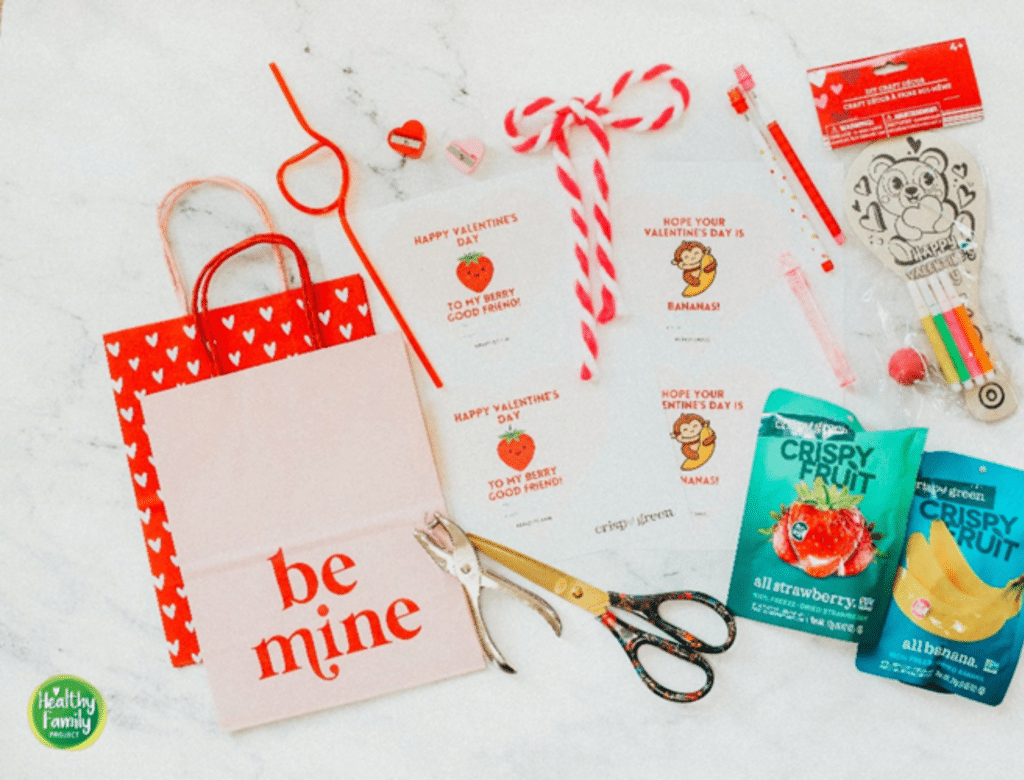Materials laid out on a table needed, including festive Valentine's Gift bags, a hole puncher, scissors, Crispy Green Crispy Fruit Strawberry and Banana snack packs, string/ribbon, Valentine's day merchandise, and Crispy Green Valentine's Day messaging.