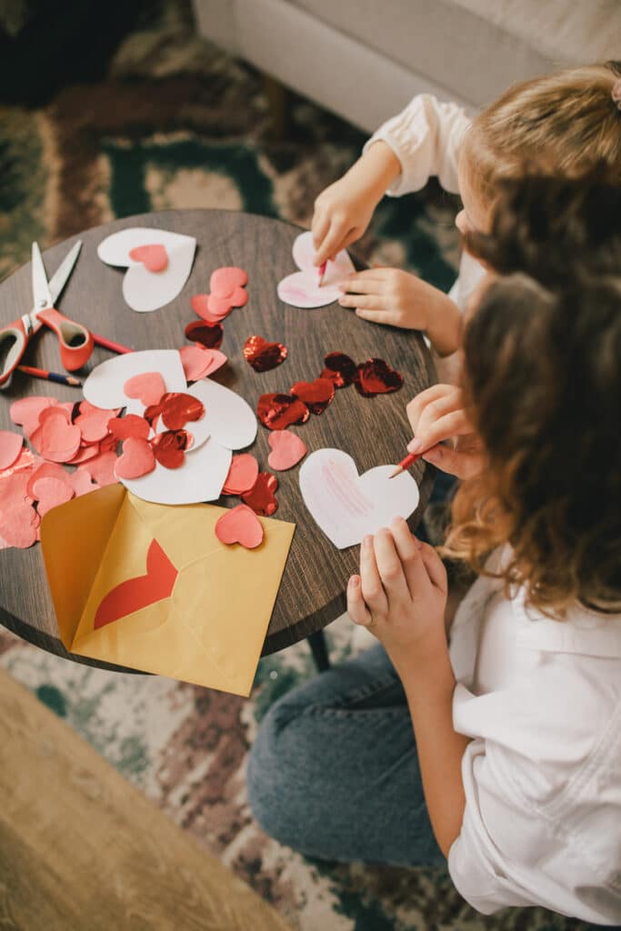 Mother and little daughter making Valentine's day cards using color paper, scissors and pencil, sitting by the table in cozy room