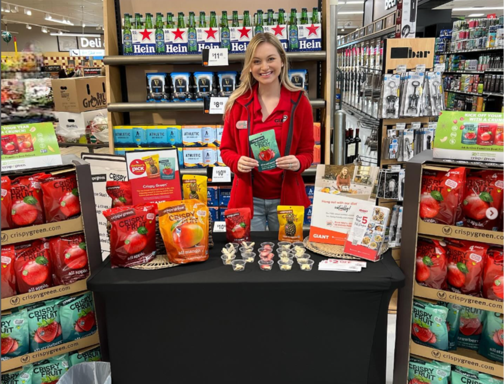 Image of a registered dietitian at a retail store, holding a bag of Crispy Fruit Strawberry. The retail dietitian is also surrounded by other Crispy Fruit multipack bags, including Mango, Apple and Pineapple. The dietitian is standing behind a black table with all the bags on it.
